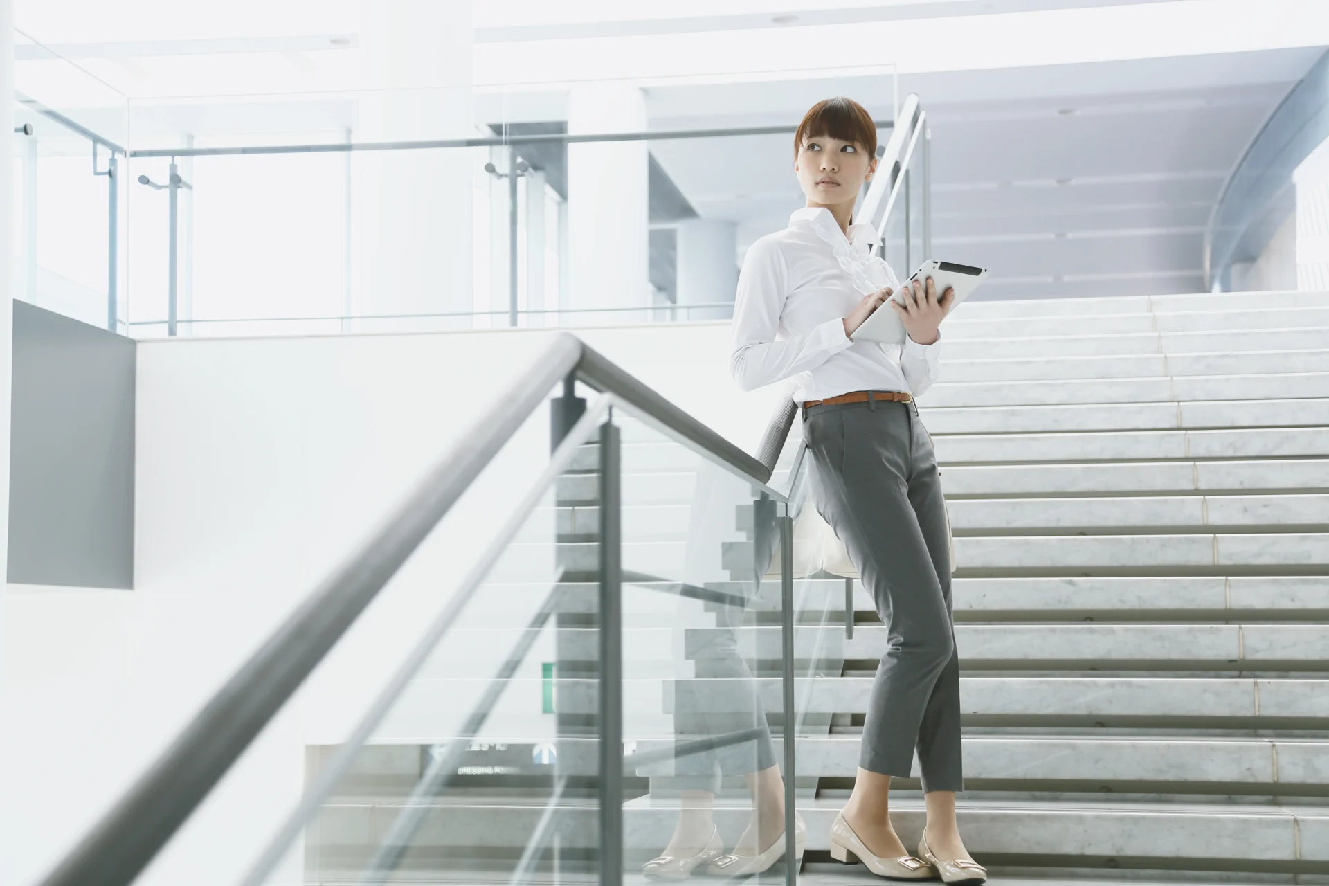 Frau auf Innentreppe eines modernen Bürogebäudes, die mit einem Tablet arbeitet, xpecto Software für Assetmanagement und Sachwerte sichtbar.